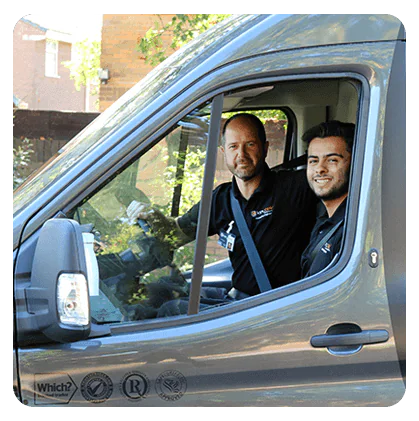 Two male locksmiths in black uniforms are seated in the front of a gray van, with one holding the steering wheel and the other wearing a seatbelt.