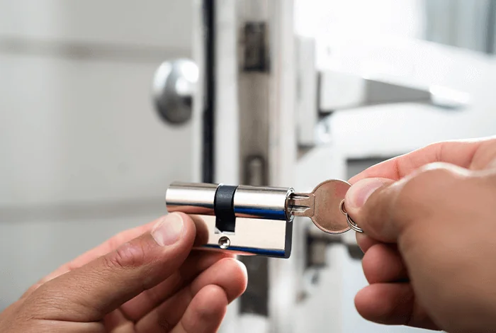 A person inserts a key into a silver door lock cylinder, preparing to unlock a door. The door handle is visible in the background.