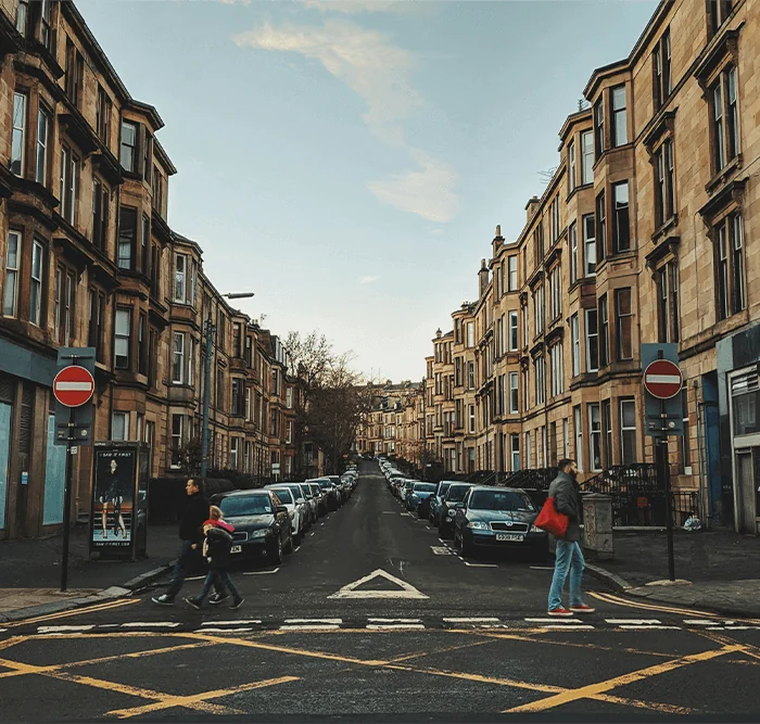 A street scene with pedestrians crossing in front of tenement buildings, flanked by parked cars on a calm day. 4 Roxburgh St, Glasgow G12 9AP, UK, Glasgow City, United Kingdom