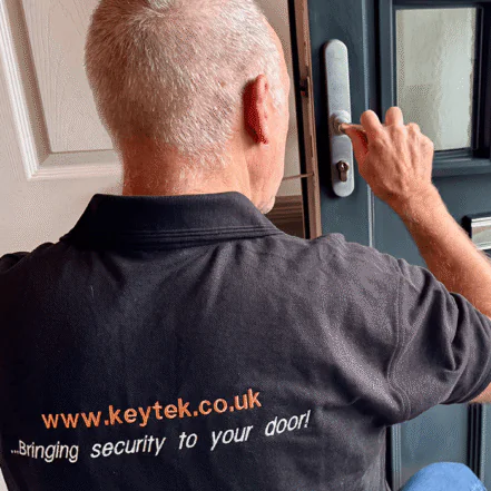 A male locksmith in a black polo shirt with "keytek" on the back is installing a door lock, focusing on enhancing home security.