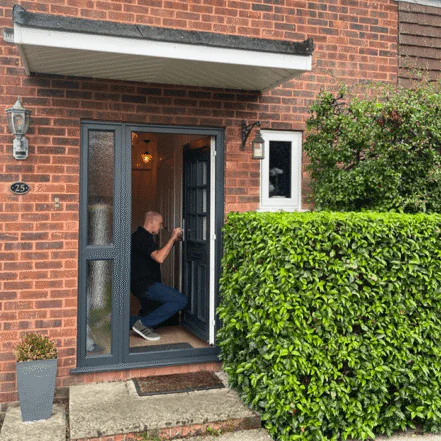 A male locksmith kneels at a front door, working on the lock, surrounded by a neatly trimmed hedge and brick wall exterior.