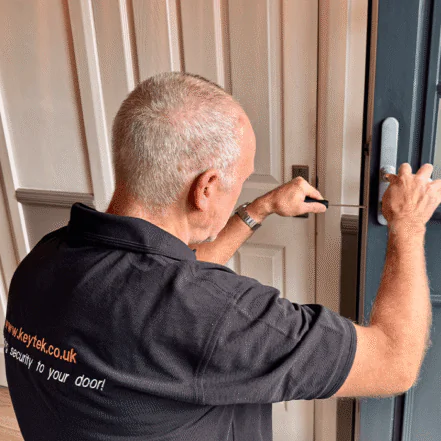 A technician wearing a black shirt from Keytek is working on securing a door lock with tools, focused on the door's mechanism.