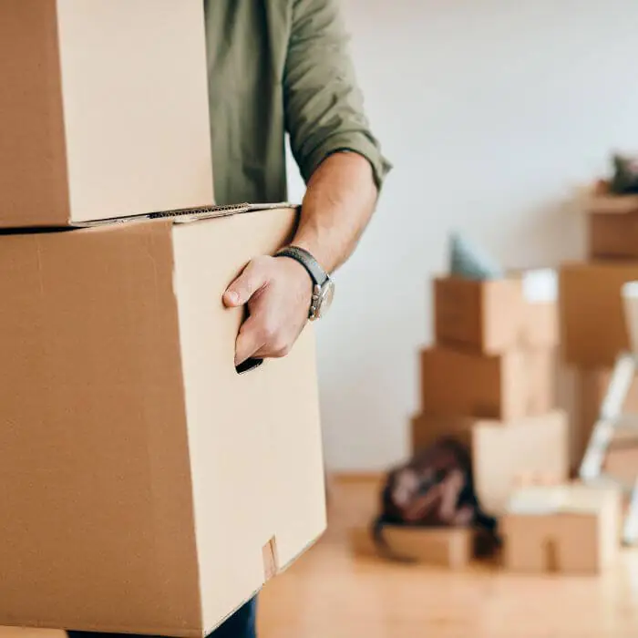 Person in an olive-green shirt carrying a tall stack of cardboard moving boxes inside a home, with additional boxes in the softly blurred background, suggesting a residential move or packing process in progress, under bright indoor lighting.