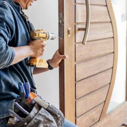 Close-up photograph of a Southeast Asian repairman in a denim jacket and light pants using a power drill on a light brown, curved wooden door with vertical paneling. He wears a tool belt and is focused on door repair in a well-lit indoor workshop or home setting.