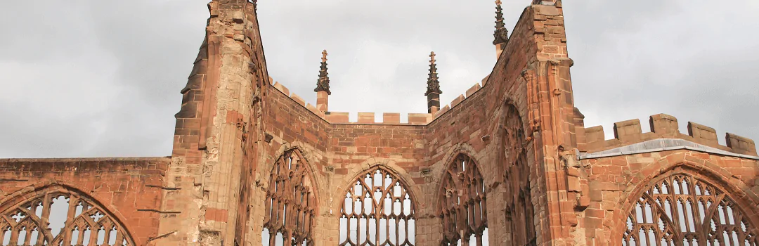 Full shot of a crumbling stone section of a historic cathedral or abbey ruin, featuring arched windows, weathered reddish-brown masonry, and remnants of stained glass under soft, overcast daylight—highlighting gothic architecture and aged craftsmanship.