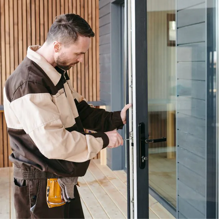 Medium shot of a handyman in a brown and beige uniform installing a uPVC glass door, using a screwdriver on the dark grey frame while standing on a wooden porch, with tool belt, gloves, and black handle and lock visible under soft natural light.