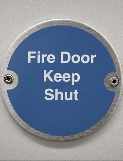 Circular sign mounted on a wall, stating "Fire Door Keep Shut" in bold white letters on a blue background.