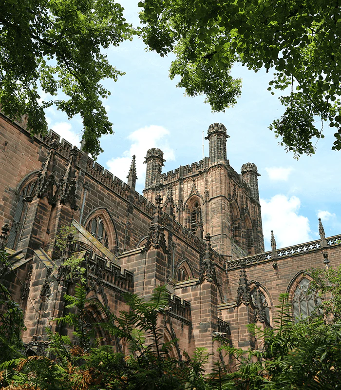 A gothic-style cathedral rises against a blue sky, framed by lush green foliage, showcasing intricate stonework and towering spires.
