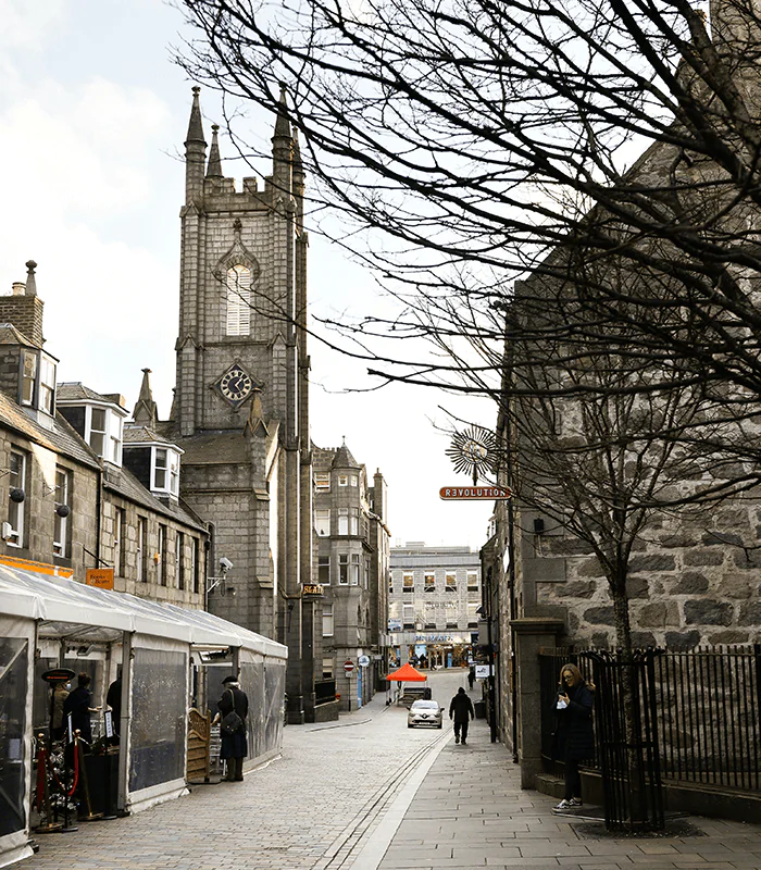 Historic street in a city showing a tall clock tower, shops, and pedestrians amid bare trees, creating a charming urban scene.