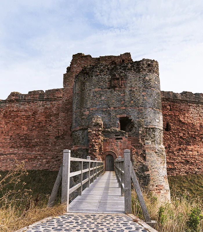 A weathered stone castle tower with a wooden bridge leading to its entrance, surrounded by lush grass and a cloudy sky.
