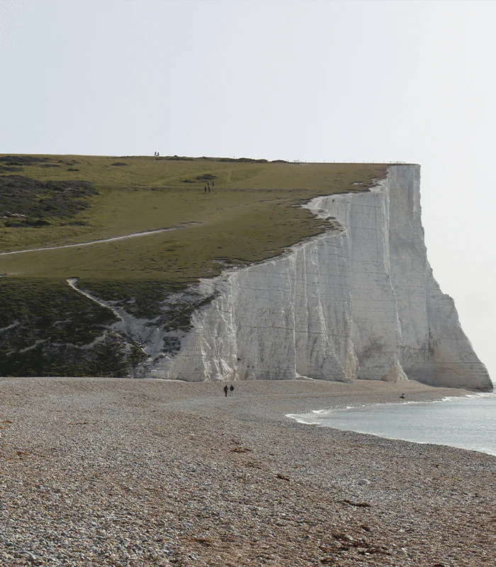 Two people walk along a pebble beach beneath towering white chalk cliffs, with lush green hills in the background and a calm sea in front.