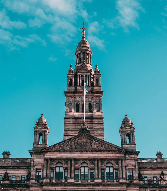 The ornate tower of a historic building rises against a bright blue sky, featuring a flag and detailed architectural elements.