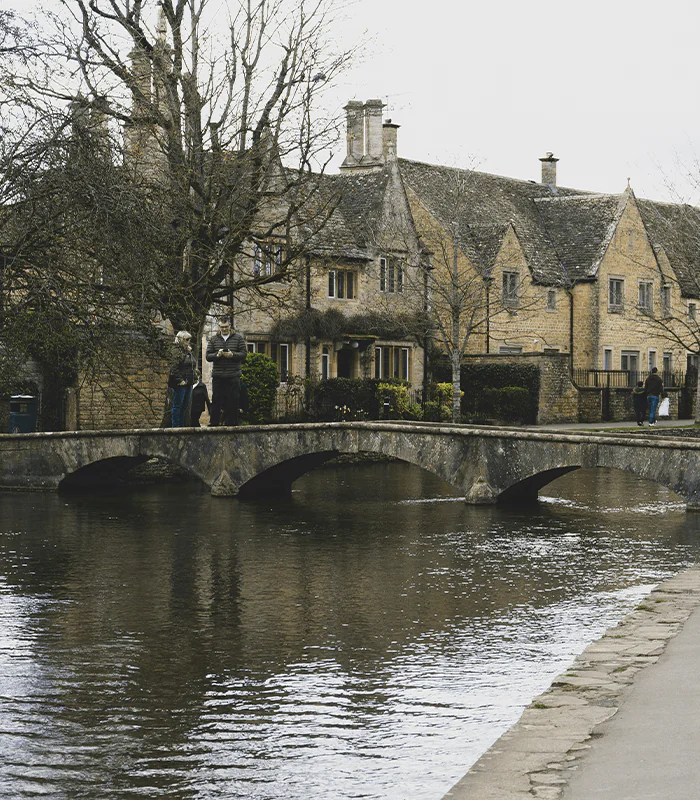 A stone bridge over a calm river, with two people standing on it; quaint Cotswold houses and leafless trees in the background.