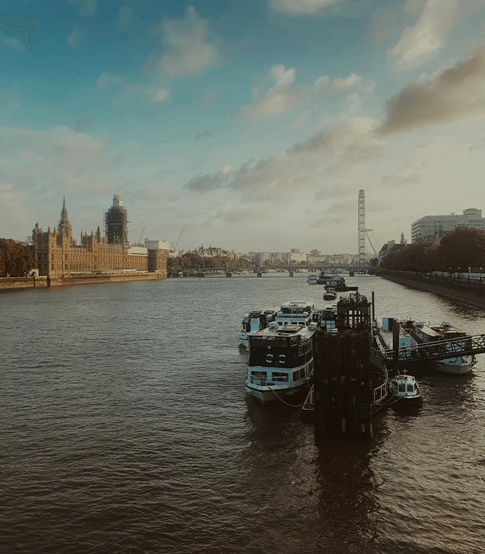 A view of the Thames River with boats docked, the Houses of Parliament and London Eye in the background under a partly cloudy sky.