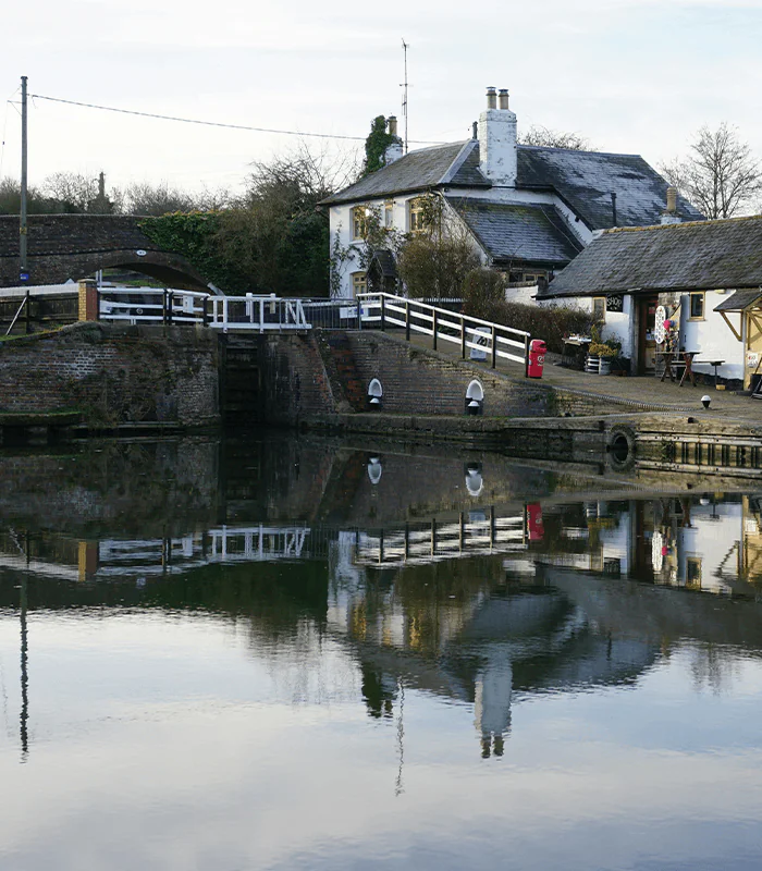 A tranquil canal scene featuring a white cottage with reflections in the water, surrounded by green trees and a wooden bridge.