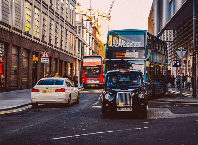 A busy urban liverpool street scene featuring a classic black taxi, a blue bus, and a white car amidst modern buildings and pedestrians.