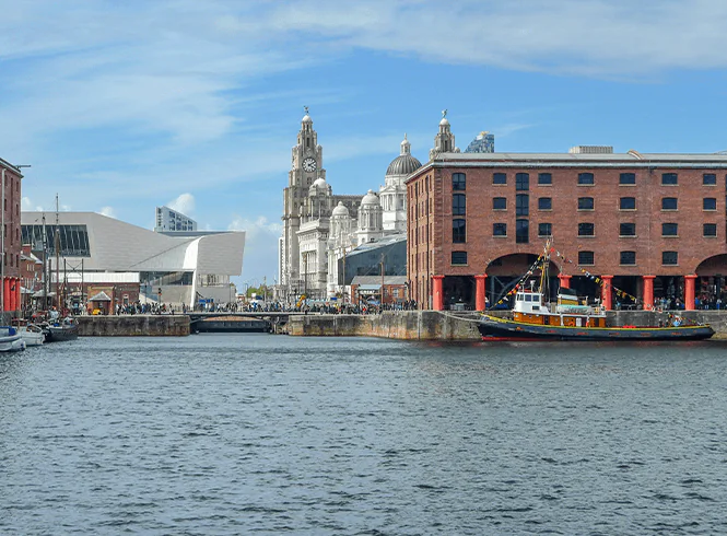 View of Liverpool's waterfront featuring historic buildings, a docked boat, and a lively crowd enjoying a sunny day.
