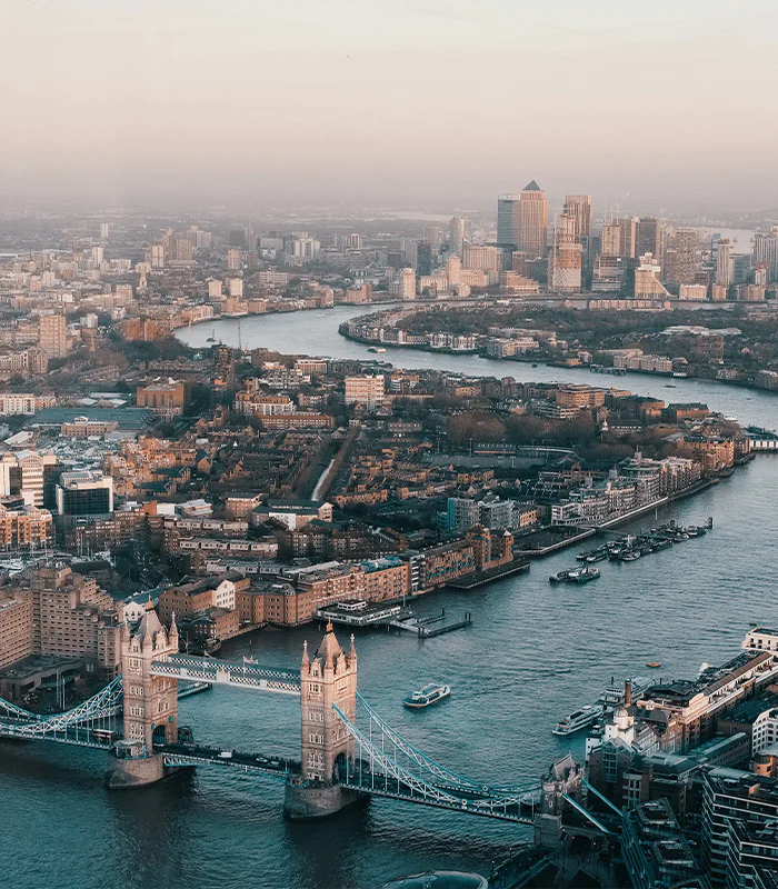 Aerial view of London featuring Tower Bridge, the River Thames, and city skyline with buildings in the background and boats on the water.