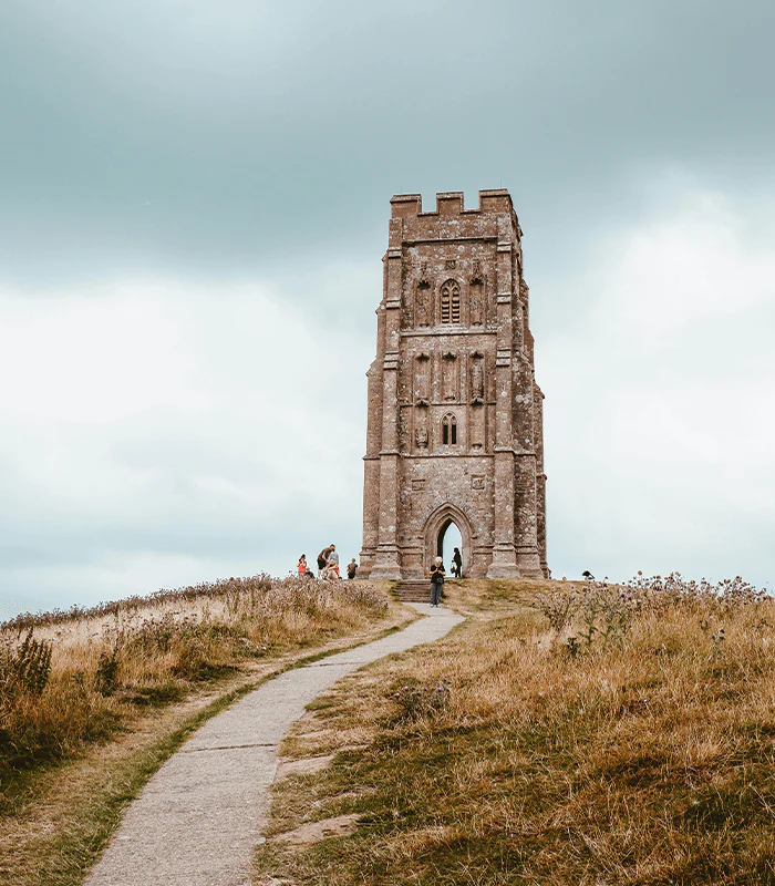A tall, stone tower stands on a grassy hill under a cloudy sky, with visitors walking along a winding path toward it.