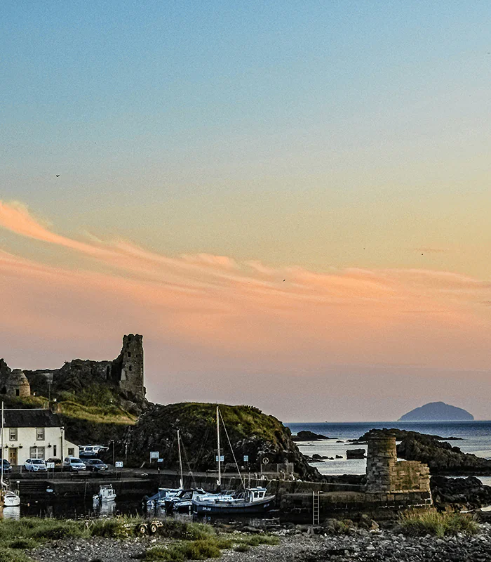 A scenic coastal view in Dunure, Ayr, featuring an ancient castle ruin, boats in a harbour, and a distant island at sunset with colourful clouds.