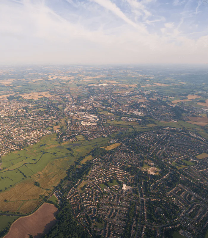 Aerial view of a sprawling suburban landscape, featuring houses, greenery, and open fields under a clear blue sky.