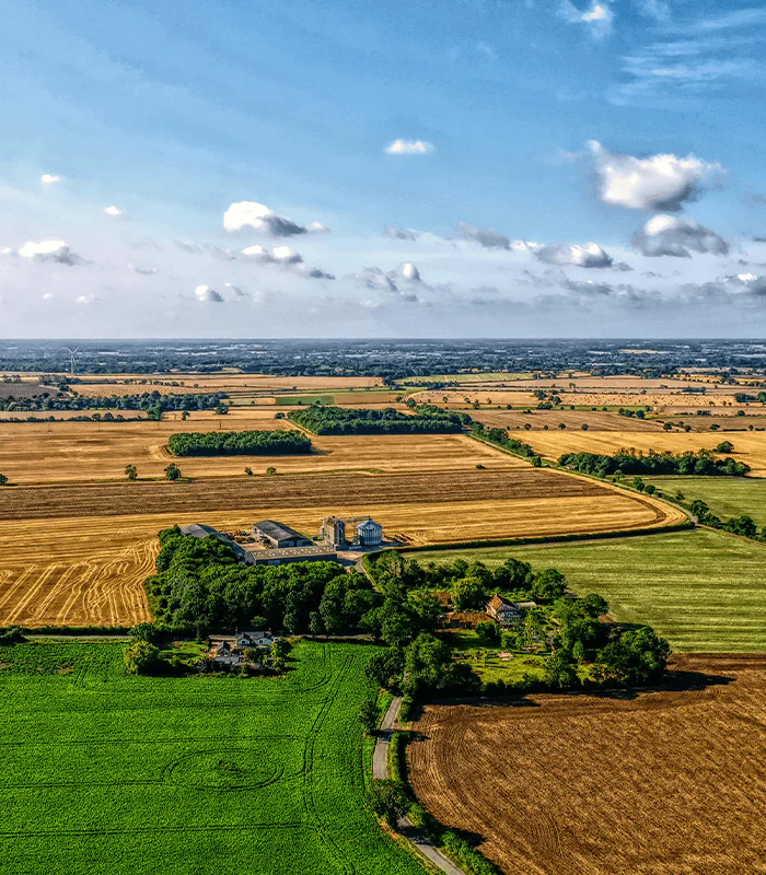 Aerial view of rural landscape in Suffolk featuring fields of varying colours, a farmstead, and scattered trees under a blue sky with clouds.