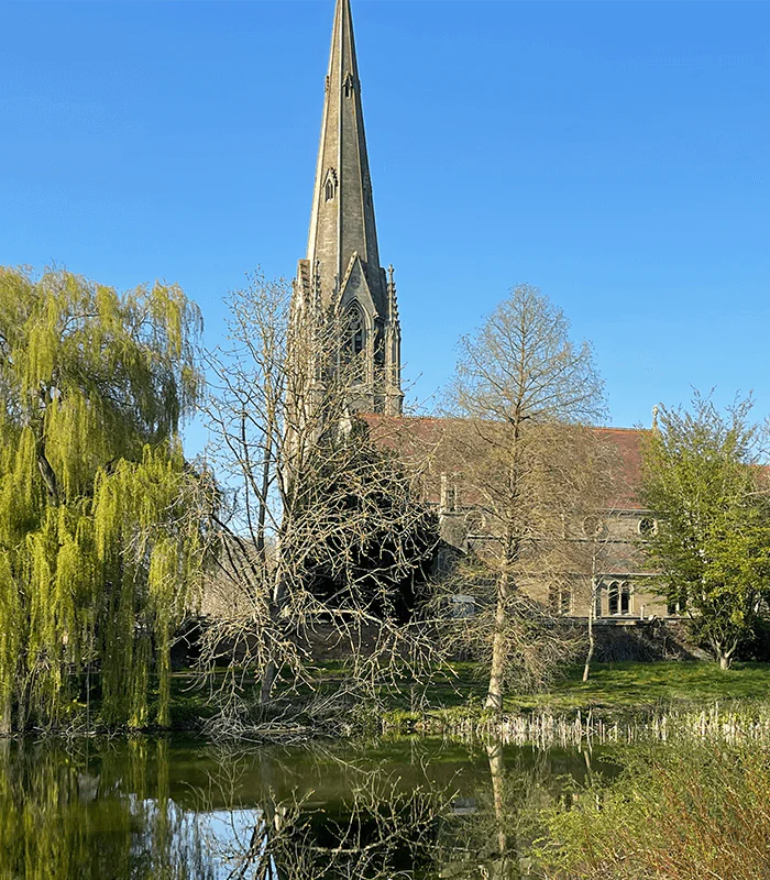 A picturesque church with a tall spire stands behind leafless trees, next to a reflective pond. Lush greenery and a clear blue sky enhance the serene scene.