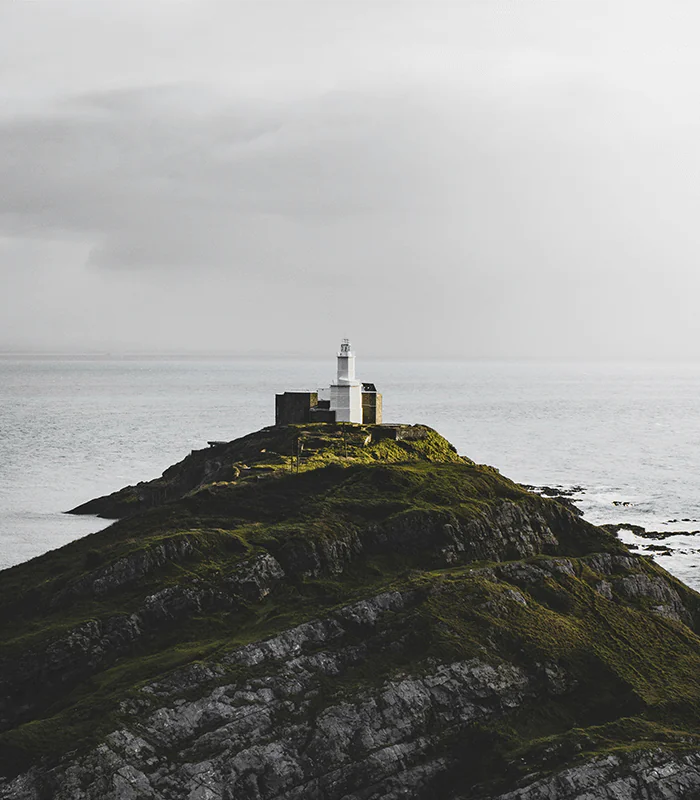 A lone white lighthouse stands atop a rugged, grassy hill beside a calm, vast ocean under a moody sky, evoking solitude and tranquillity.