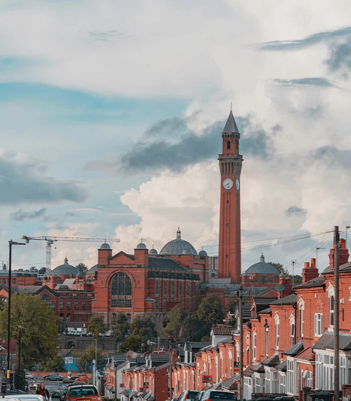 A tall clock tower and red-brick buildings dominate the skyline under a partly cloudy sky. The foreground features cars and terraced houses lining a sloped street.