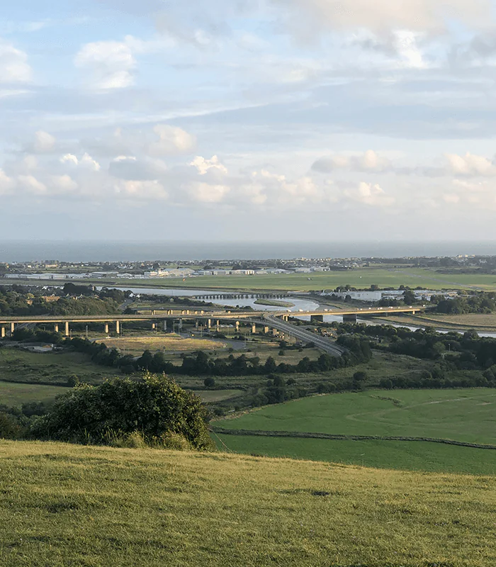 A scenic landscape with rolling green hills in the foreground. A series of bridges spans across a river, leading to a town under a cloudy blue sky.