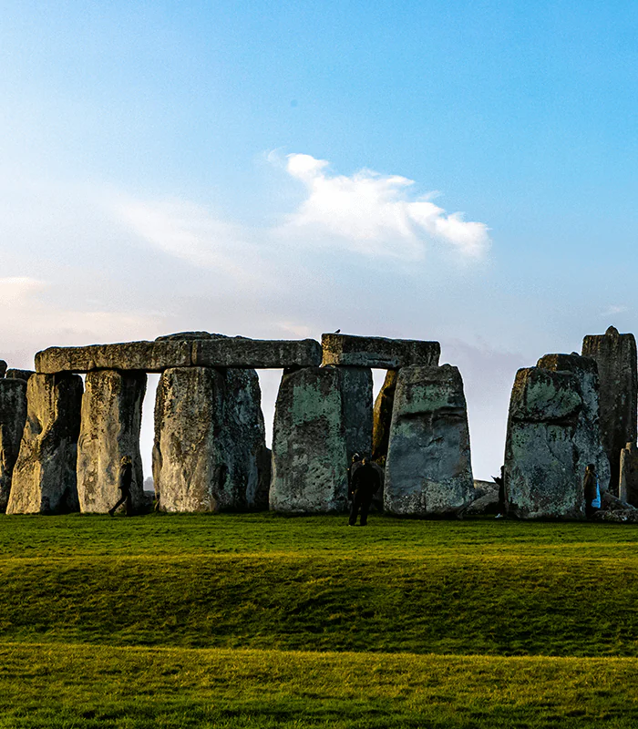 Stonehenge at sunset with a clear blue sky. Large stone slabs form a circular structure on green grass. Visitors walk around, evoking a sense of wonder.