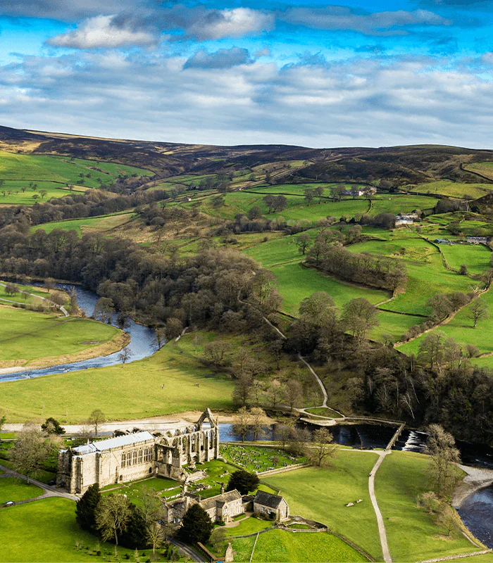 Aerial view of lush green Yorkshire countryside with rolling hills. A historic stone abbey sits near a winding river, under a partly cloudy blue sky. Serene and picturesque.