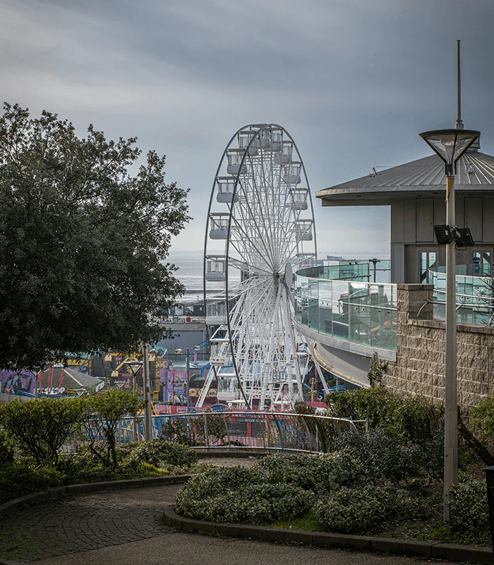 A white ferris wheel stands in the distance, framed by greenery and overlooking a colorful amusement park near the coast.