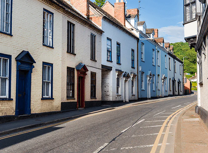 A picturesque street lined with colorful houses in various pastel shades, basking in sunlight with a clear blue sky and greenery in the background.