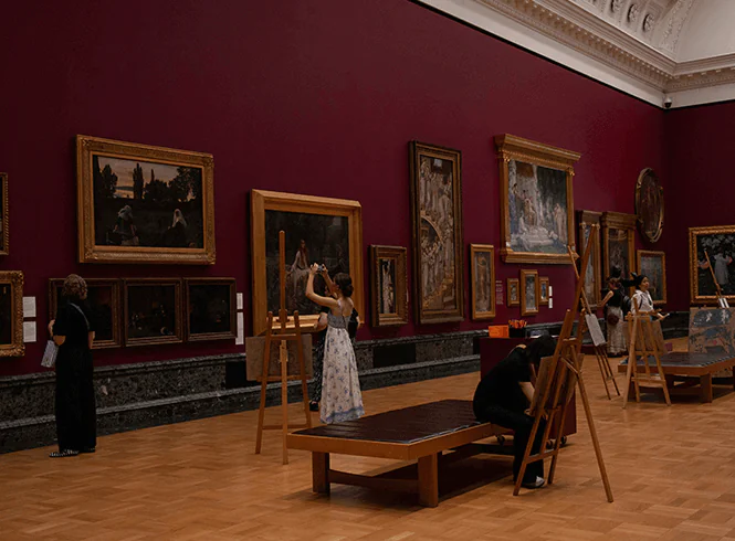 Visitors admire artwork in a gallery with red walls, while some sketch on easels. Richly framed paintings adorn the space.