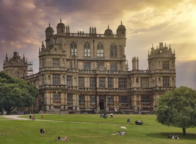 Wollaton Hall in Nottingham, UK, a grand multi-tiered stone mansion with towers and ornate windows, bathed in warm sunset light against a dramatic sky, overlooking a sloping green lawn where people picnic, walk, and enjoy the historic parkland setting.