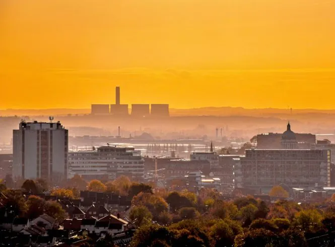 NOT 3 Panoramic view of Nottingham cityscape at sunrise or sunset with a golden sky, autumn trees in the foreground, rows of dark-roofed houses, a central high-rise, domed building, and distant industrial structures with tall chimney, softened by atmospheric haze