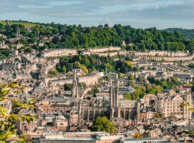 Panoramic view of Bath, England, featuring the grand Gothic Bath Abbey with soaring spires surrounded by rows of light stone Georgian terraces, rolling green hills, and lush trees beneath a softly clouded sky.