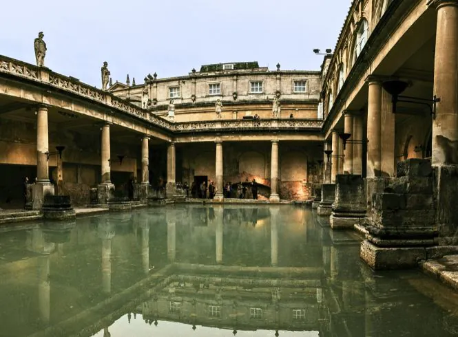 Historic view of the Roman Baths in Bath, England, featuring the steaming Great Bath with green mineral water surrounded by weathered stone columns, statues of Roman figures, and warm ancient architecture under soft natural light.