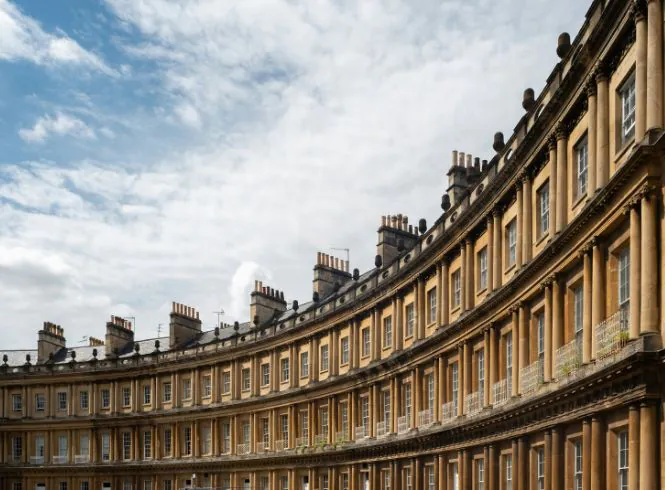 Elegant view of the Royal Crescent in Bath, England, showing a sweeping curve of Georgian terraced townhouses built from warm honey-colored Bath stone, with tall sash windows, chimneys, and a soft partly cloudy sky above.