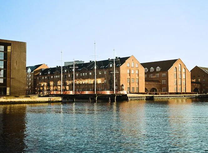 Serene waterfront view at Gloucester Docks with historic red-brick warehouse buildings reflected in calm water, tall narrow windows and pitched roofs hinting at their industrial past, under a clear blue sky with late afternoon light
