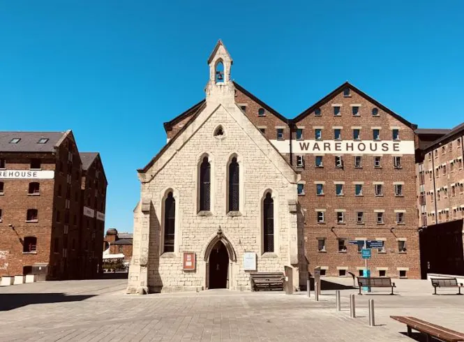 Tranquil view of Mariners Church in Gloucester, a historic light stone building with Gothic features and a small spire, standing in a sunlit paved square beside large red-brick warehouses marked ‘WAREHOUSE,’ under a vivid blue sky