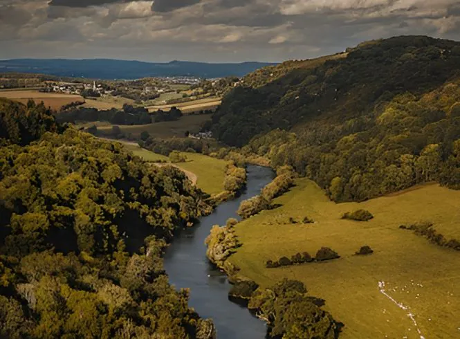 Panoramic view of a winding river in Gloucester, Gloucestershire, flowing through a lush green valley with forested hills, open fields, and partly cloudy skies casting light and shadow across the serene landscape.