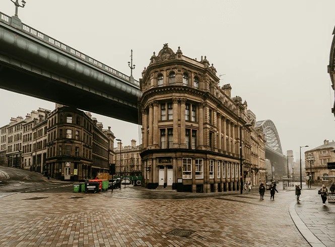 Street scene in Newcastle upon Tyne, England, with people walking along Grey Street, renowned for its Georgian architecture. The Tyne Bridge spans the River Tyne in the background, while the High Level Bridge crosses above the street