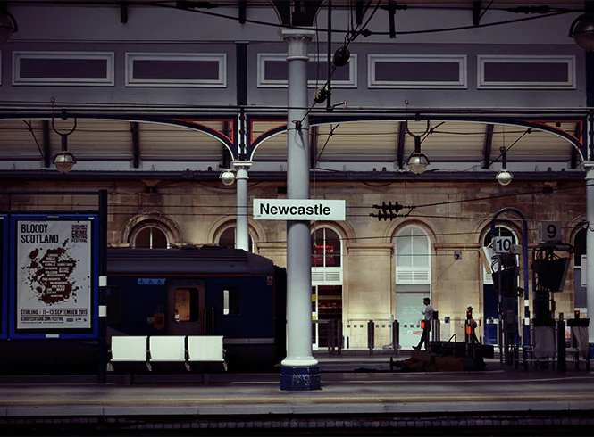 Newcastle Central Station platform with a train and a grey post displaying 'Newcastle' signage. The historic station in North East England connects to major UK cities and the local Metro. A poster for the 'Bloody Scotland' festival from September 2015 is visible on the platform.