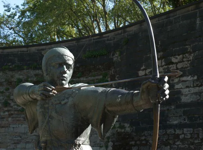 Bronze statue of Robin Hood in Nottingham, England, showing the legendary archer mid-draw with bow and arrow, wearing a hooded tunic, set on a stone-paved area against a weathered brick wall with mounted plaques, highlighting historic heritage.