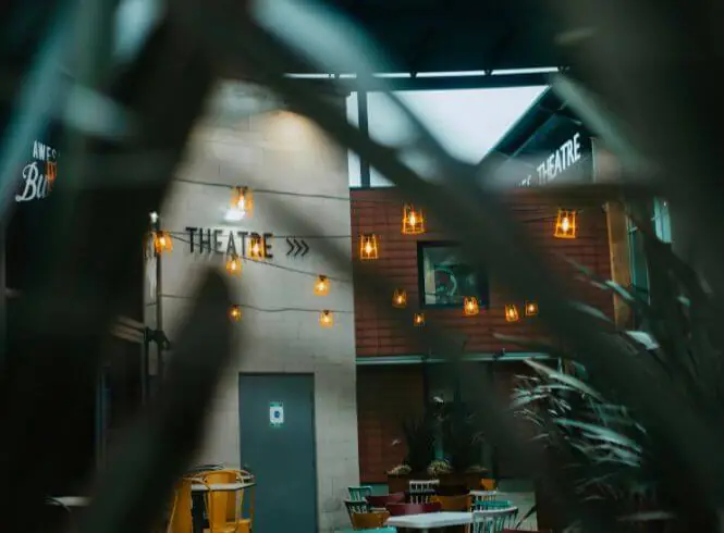 Milton Keynes theatre interior softly framed by green leaves, with warm ambient lighting from string lights and wall lamps creating a cozy atmosphere. The word ‘THEATRE’ with directional arrows is visible on the wall, guiding visitors toward a performance space. Modern seating and tables sit beneath the golden glow, blending natural elements with contemporary design to evoke creativity, culture, and community