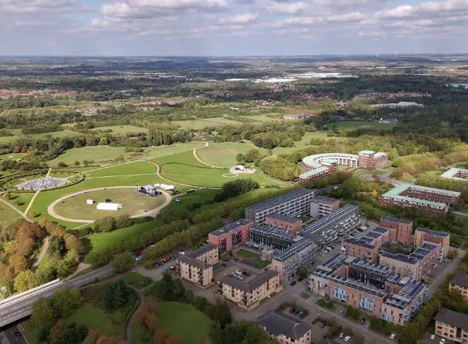 Aerial view over Avebury Boulevard in Milton Keynes looking towards Campbell Park and contemporary art installation. The image showcases manicured green spaces, winding pathways, modern brick buildings with solar panels, and a distant forest under a partly cloudy sky. The scene highlights the balance between urban design, public parks, and natural landscapes in Milton Keynes