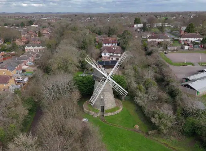 Aerial view of Bradwell Windmill in Milton Keynes, Buckinghamshire, England, showing a historic gray stone windmill with white sails surrounded by bare trees, green grass, nearby residential houses with red-tiled roofs, and a partly overcast sky on a cloudy day, highlighting the contrast between the historic structure and the modern landscape