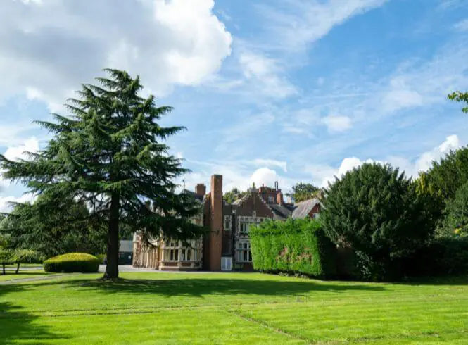 Bletchley Park on Sherwood Drive in Bletchley, Milton Keynes, UK, showcasing a stately multi-story red-brick mansion surrounded by lush green lawns, mature trees, and neatly trimmed hedges under a bright, partly cloudy sky. The image highlights classic architecture, landscaped grounds, and the serene, elegant setting of the historic property.